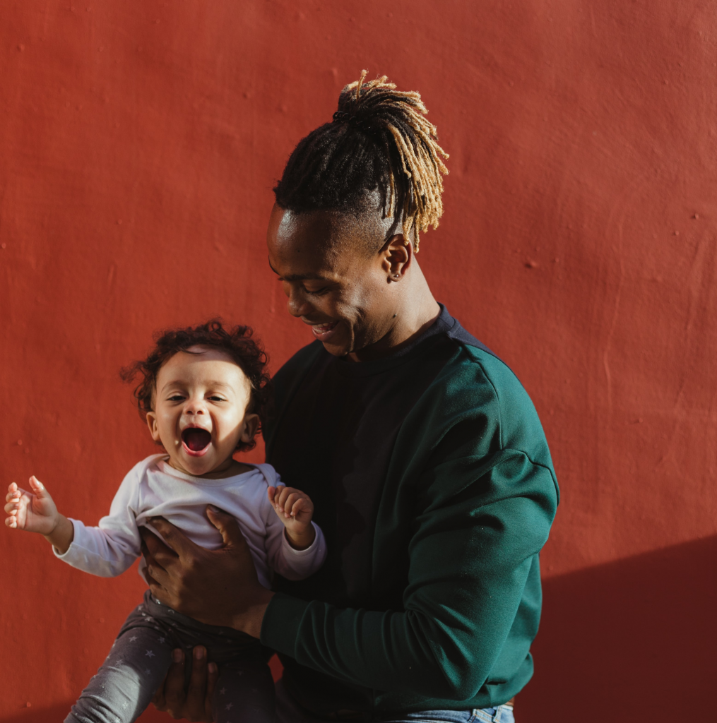 A young black man holds his toddler daughter. He's laughing and smiling at her. She's happy and smiling too. 