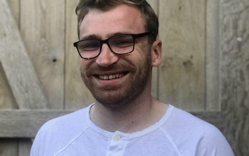 A man with short hair wearing glasses and a white tshirt smiles at the camera.