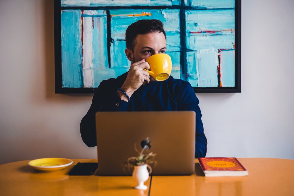 a man sits at a desk with a laptop in front of him. He's sipping coffee from a large yellow mug. 