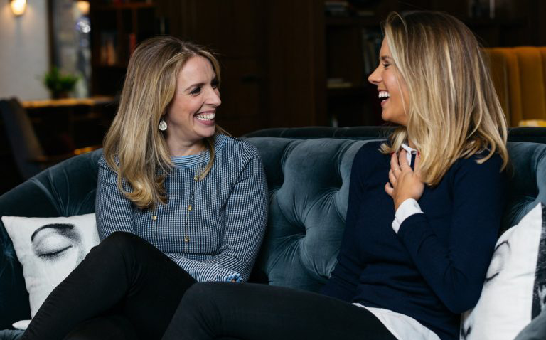 Two blonde women sit on a blue sofa. They're looking at each other and laughing