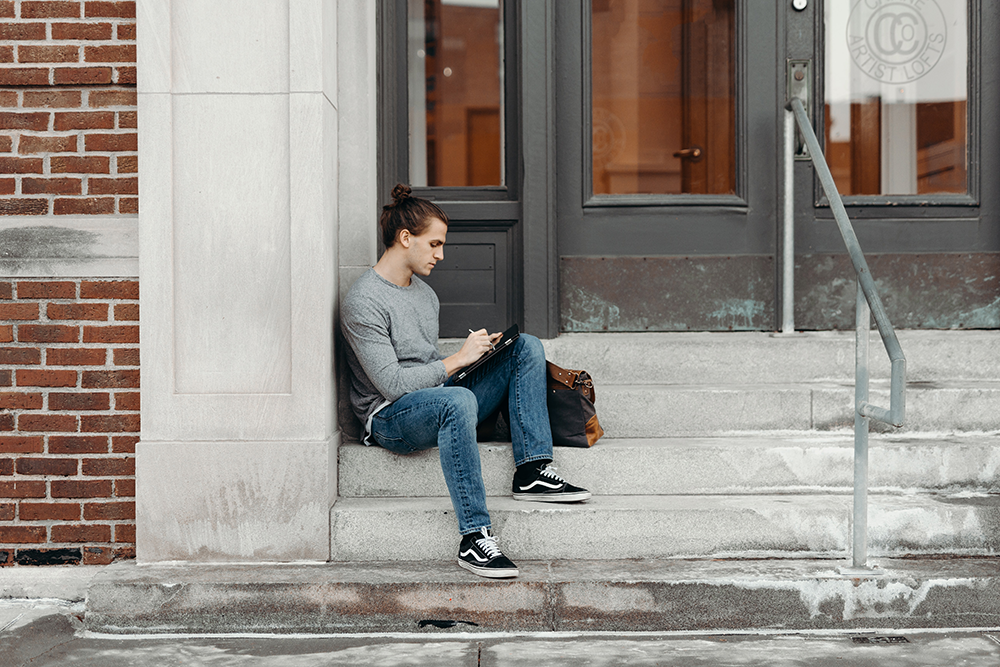 A man sits on the steps in front of a building. He's working on an ipad. He's wearing jeans, black and white Vans trainers and a grey long sleeved top. 